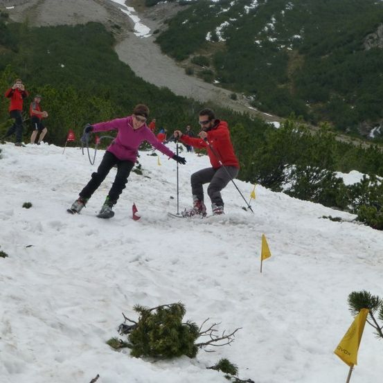 Zwei Skifahrer navigieren einen schneebedeckten Berghang, mit Fahnen, die den Kurs markieren. Zuschauer beobachten von hinten.