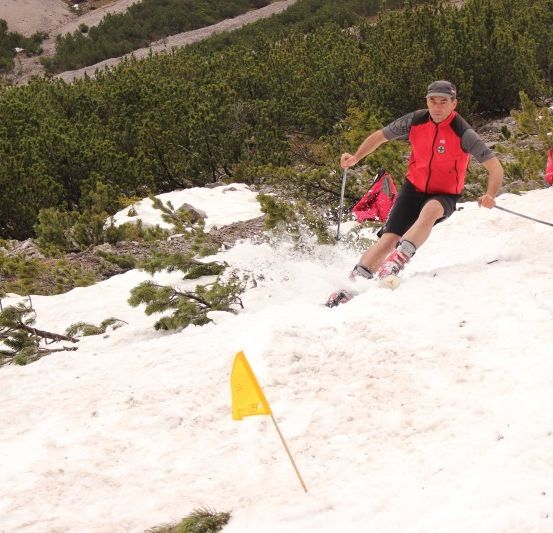 Ein Mann, der einen schneebedeckten Berg hinunterskit. Er trägt eine rote Weste und schwarze Shorts. Er hält Skistöcke und passiert eine gelbe Flagge.