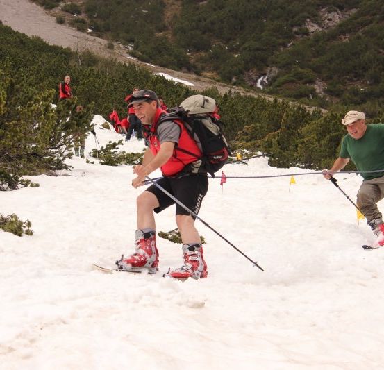 Eine Gruppe von Menschen skifahrer auf einem verschneiten Berg. Ein Mann trägt einen roten Weste und einen Rucksack. Ein anderer Mann hält eine Seil.