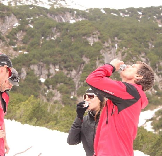 Drei Personen in roten Jacken trinken Wasser in einem verschneiten Gebirgsgebiet.
