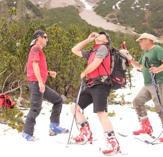 Drei Männer in Skiausrüstung, einer trinkt aus einer Flasche, stehen im Schnee mit Bäumen und Bergen im Hintergrund.