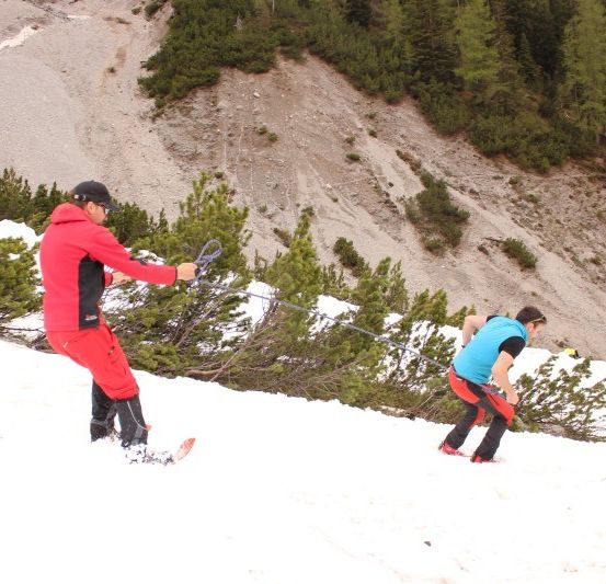 Zwei Skifahrer auf einem verschneiten Berg, einer zieht den anderen mit einem Seil.