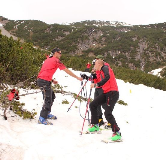 Drei Skifahrer in roten Jacken stehen auf einem verschneiten Berg, einer hält Skistöcke und ein anderer eine Kamera.