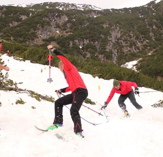 Drei Skifahrer in roten Jacken und schwarzen Hosen fahren einen verschneiten Berg hinunter. Ein Skifahrer hebt den Arm.