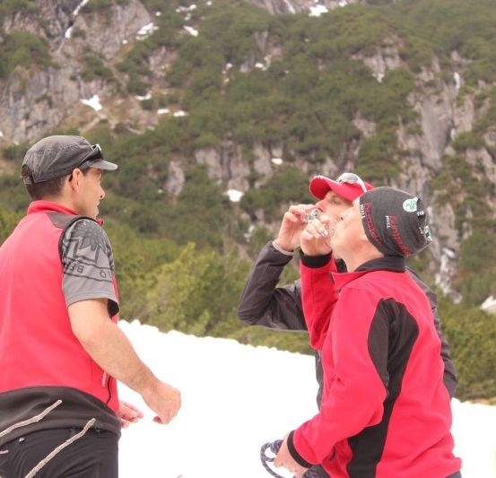 Drei Männer in roten Jacken stehen auf einem verschneiten Berg, einer trinkt aus einer Flasche.