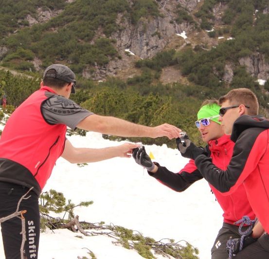 Drei Männer in roten Jacken stehen auf einem verschneiten Berg, einer hält eine Kamera.