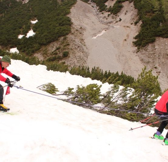 Zwei Skifahrer steigen einen schneebedeckten Hang hinauf, einer zieht den anderen mit einem Seil. Sie tragen beide rote Jacken und schwarze Hosen.