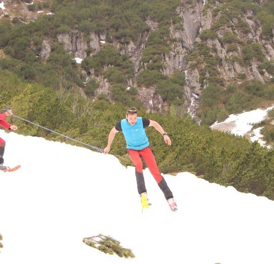 Zwei Personen skifahren bergauf auf einem verschneiten Berg, eine zieht die andere mit einem Seil.