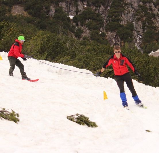 Zwei Personen steigen auf einem verschneiten Berg ab, einer zieht den anderen mit einem Seil. Sie tragen rote Jacken und schwarze Hosen.
