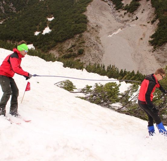 Zwei Skifahrer steigen einen schneebedeckten Hang hinauf, einer hält eine Seil, während der andere hinterher schippert.