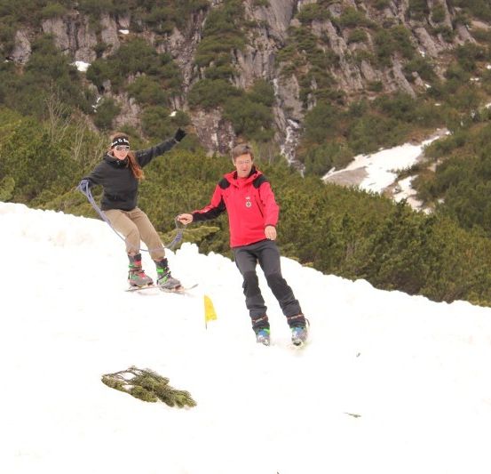 Zwei Personen steigen einen schneebedeckten Berghang hinunter. Die Person vorne trägt eine schwarze Jacke und Hose, während die Person dahinter eine rote Jacke und Hose trägt. Sie halten sich an einem Seil fest.