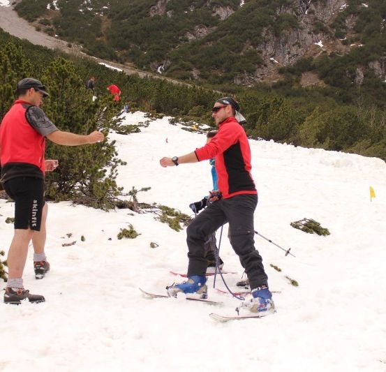 Zwei Männer in roter und schwarzer Skiausrüstung stehen auf einem verschneiten Berg, einer hält einen Skistock.