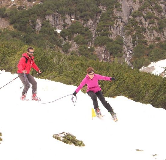 Zwei Skifahrer, einer in Rot und einer in Pink, fahren mit einem verbindenden Seil einen schneebedeckten Berg hinunter.