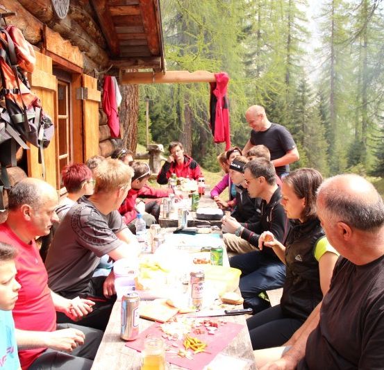 Eine Gruppe von Menschen sitzt um einen Tisch vor einer Holzhütte, isst und trinkt. Einige tragen Jacken und Mützen. Bäume und ein Rasenbereich sind im Hintergrund zu sehen.