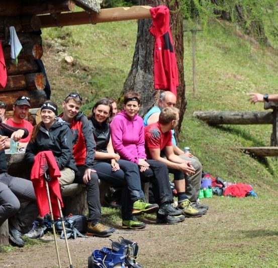 Eine Gruppe von Wanderern in Outdoor-Kleidung sitzt auf einer Bank im Wald. Ein Mann in einem roten Shirt steht und spricht mit ihnen.