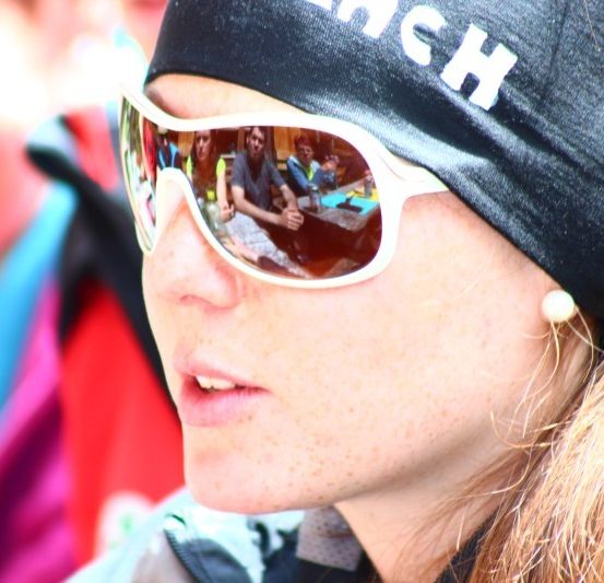 Eine Frau mit Stirnband und Sonnenbrille, möglicherweise bei einer Skiveranstaltung, mit einem verschwommenen Hintergrund, der Menschen an einem Tisch zeigt.