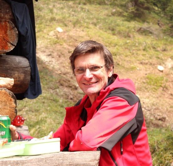 Ein Mann in einem roten Mantel sitzt an einem Holztisch im Freien, lächelt und hält eine grüne Dose. Hinter ihm sind Holzscheite und ein blauer Stoff gestapelt.