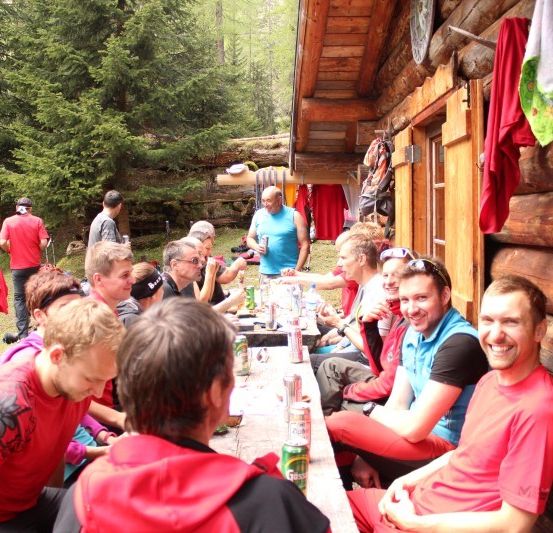 Eine Gruppe von Menschen in roten und blauen Shirts sitzt um einen langen Tisch vor einer Blockhütte, lächelt und trinkt.