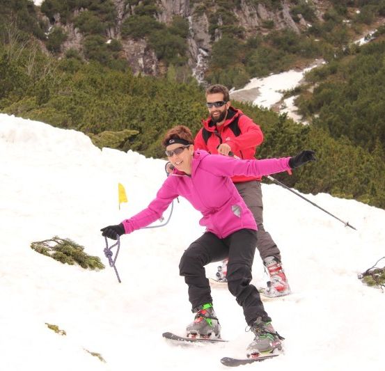 Eine Frau in einer pinken Jacke und schwarzen Hosen skifahrer eine schneebedeckte Bergflanke hinunter, mit einem Mann in einer roten Jacke und Sonnenbrille hinter ihr.