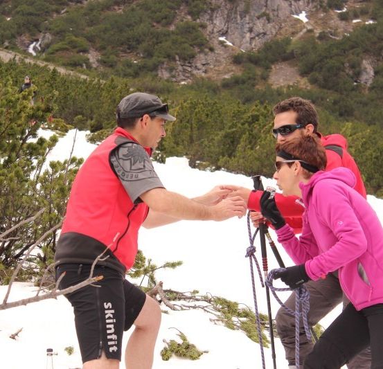 Drei Personen, in Winterkleidung gekleidet, stehen in einem verschneiten Berggebiet. Der Mann in der Mitte schüttelt der Frau rechts von ihm die Hand, während der Mann links von ihm sie ansieht.