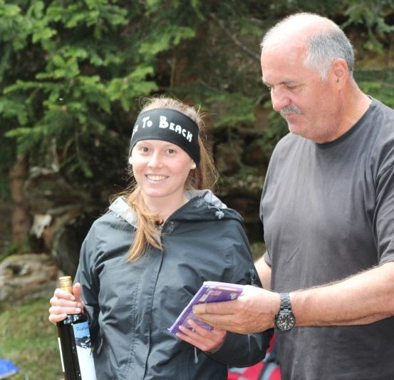 Eine Frau in einer schwarzen Kopfband und Jacke hält eine Flasche Wein, während ein Mann in einem grauen T-Shirt einen lila Umschlag hält.