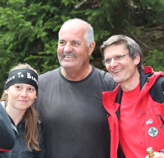 Drei Personen posieren für ein Foto im Wald. Der Mann in der Mitte hat einen Schnurrbart und trägt ein graues T-Shirt. Der Mann rechts trägt eine rote Jacke und hält eine Flasche. Die Frau links trägt eine schwarze Kopfband.
