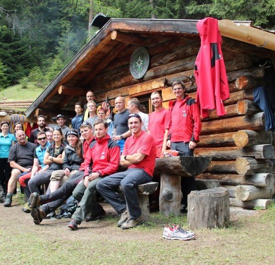 Eine Gruppe von Menschen in roten Jacken sitzt auf einer Bank vor einer Holzhütte.