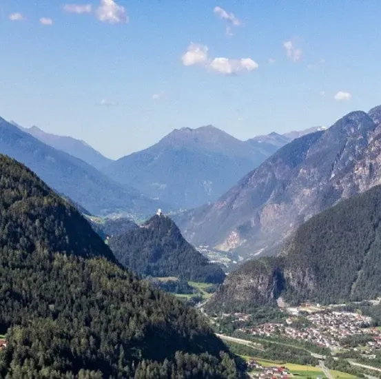 Ein atemberaubender Luftblick auf eine Bergregion mit dichten Wäldern, einem kleinen Dorf und einer Burg auf einem Hügel unter einem blauen Himmel mit verstreuten Wolken.