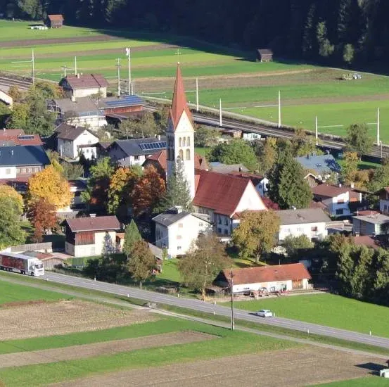 Ein Luftbild eines kleinen Dorfes mit einer Kirche, Häusern und grünen Feldern. Die Kirche hat einen Turm und ein rotes Dach. Ein LKW ist auf der Straße.
