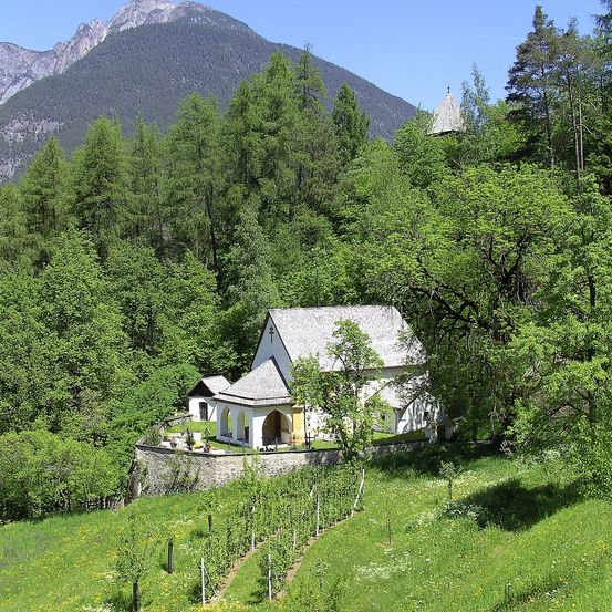 Eine kleine Kirche mit grauem Dach und gelben Wänden liegt in einem üppigen grünen Tal, umgeben von Bäumen und Bergen.
