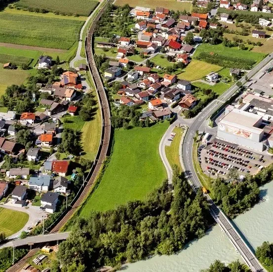 Luftaufnahme einer kleinen Stadt mit Häusern, einem Fluss und einer Straße. Die Stadt hat eine Mischung aus Wohn- und Geschäftsgebäuden, mit einem großen Gebäude auf der rechten Seite.