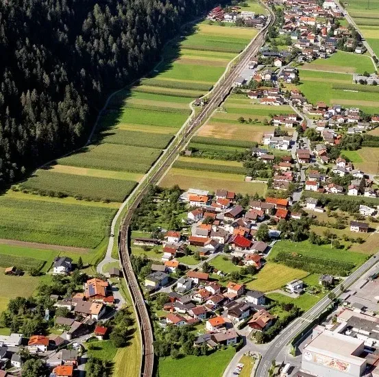 Eine Luftaufnahme einer kleinen Stadt zwischen grünen Feldern und einem Fluss, mit Häusern und einer Eisenbahnstrecke, die hindurchführt.