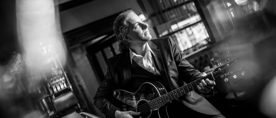 Ein Mann in Anzug spielt eine akustische Gitarre in einem Restaurant. Die Szene ist in Schwarz-Weiß gehalten.
