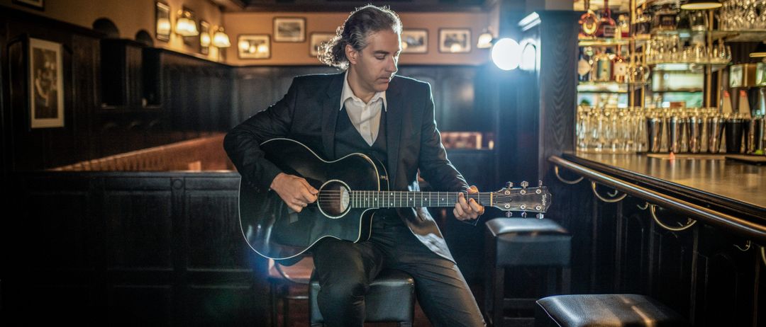 Ein Mann in einem Anzug spielt Gitarre, während er auf einem Hocker in einer Bar sitzt.