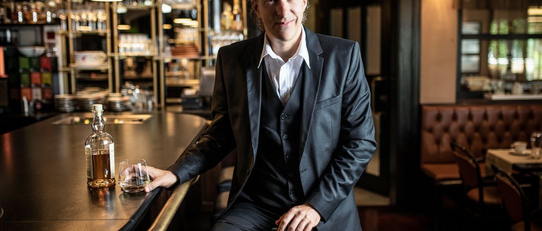 Ein Mann in einem schwarzen Anzug sitzt an einer Bar, lächelt und hält eine Gitarre. Hinter ihm befindet sich eine Bartheke mit einer Flasche Whiskey und einem Glas. Im Hintergrund sind Regale mit Gläsern und Flaschen zu sehen.