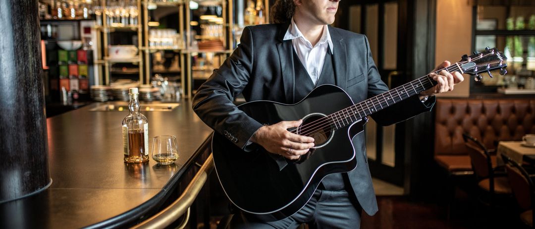 Ein Mann in einem Anzug spielt eine Gitarre in einer Bar. Eine Flasche und zwei Gläser stehen auf der Theke.