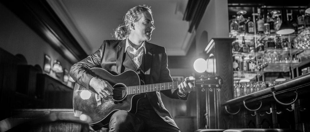 Ein Schwarz-Weiß-Foto eines Mannes in Anzug, der auf einem Hocker in einem Restaurant sitzt und eine Gitarre spielt.