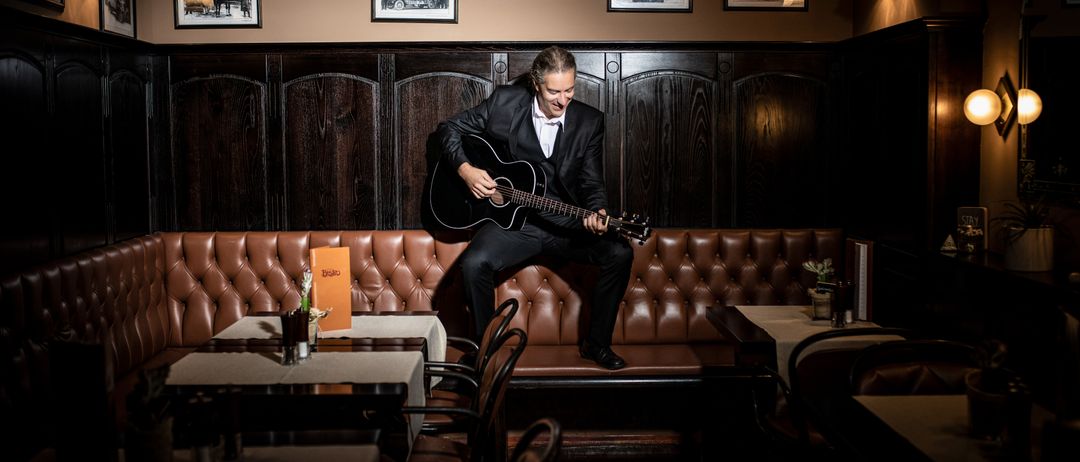 Ein Mann in Anzug spielt Gitarre, während er auf einer Bank in einem schwach beleuchteten Restaurant sitzt.