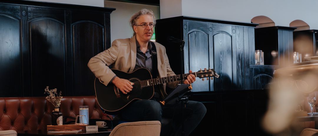 Ein Mann spielt Gitarre auf einem Hocker in einem Raum mit Stühlen und einem Tisch. Der Raum hat dunkle Holzschränke, eine weiße Wand und einen Teppichboden.