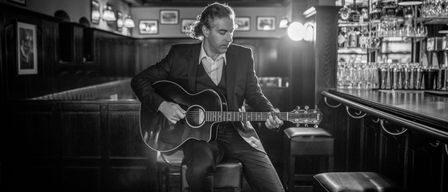 Ein Mann in einem Anzug sitzt auf einem Hocker und spielt eine akustische Gitarre in einer schwach beleuchteten Bar.