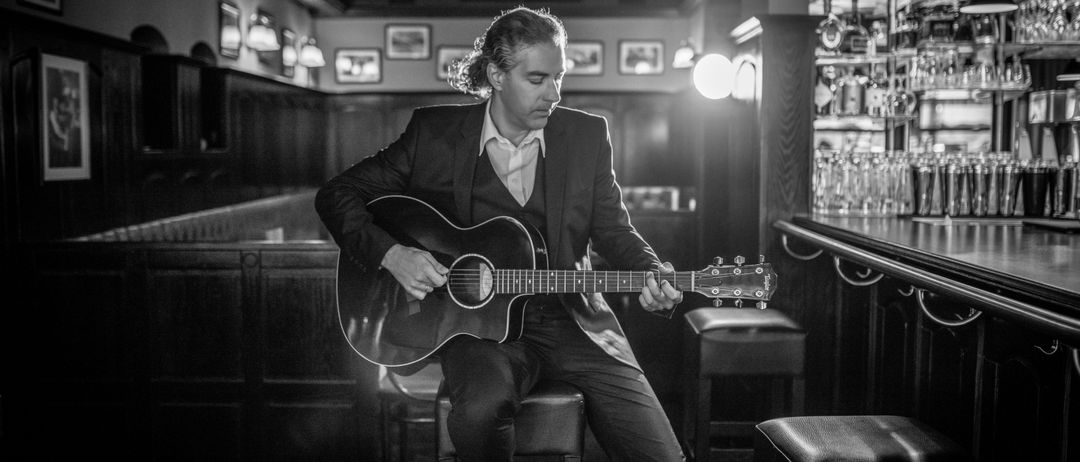 Ein Mann in einem Anzug sitzt auf einem Hocker und spielt eine akustische Gitarre in einer schwach beleuchteten Bar.