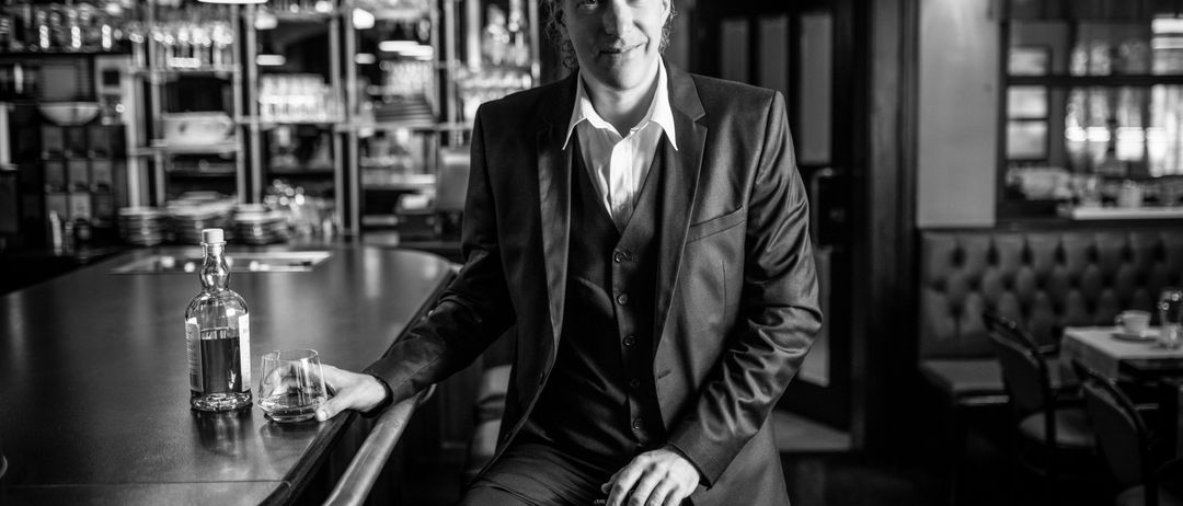 Ein Mann sitzt mit einer Gitarre auf einem Barhocker, lächelt, ein Glas und eine Flasche in der Nähe. Dahinter steht eine Bar mit Regalen voller Gläser.