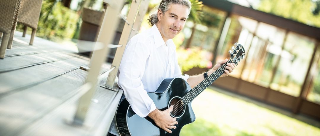 Ein Mann in einem weißen Hemd und Jeans steht neben einer Metallbrüstung und hält eine schwarze akustische Gitarre. Dahinter ist ein Gebäude mit Glaswänden und einem grünen Rasen zu sehen.