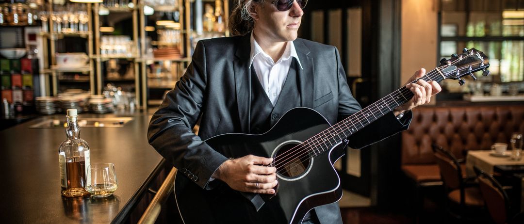 Ein Mann in einem Anzug spielt eine schwarze Gitarre in einer Bar mit Regalen und einem Glas dahinter.