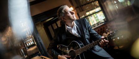 Ein Mann mit einer Gitarre sitzt in einem Raum, wahrscheinlich in einem Restaurant, und schaut nach oben. Er trägt einen schwarzen Anzug und spielt die Gitarre.