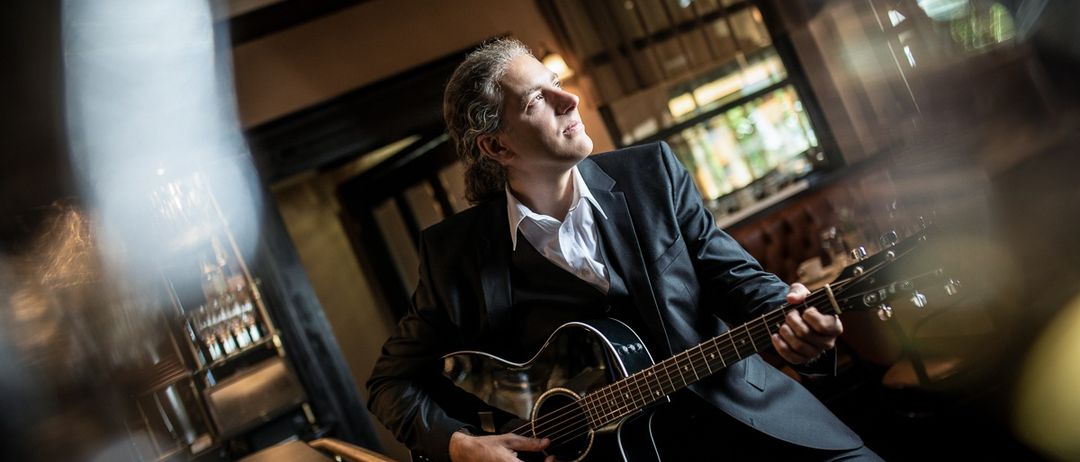 Ein Mann mit einer Gitarre sitzt in einem Raum, wahrscheinlich in einem Restaurant, und schaut nach oben. Er trägt einen schwarzen Anzug und spielt die Gitarre.