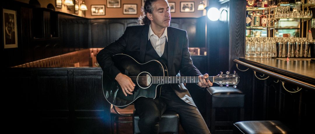 Ein Mann in einem Anzug sitzt auf einem Hocker und spielt eine Gitarre in einer schwach beleuchteten Bar. Die Bar ist mit dunklen Holzpaneelen und einer Bartheke mit Flaschen ausgestattet.