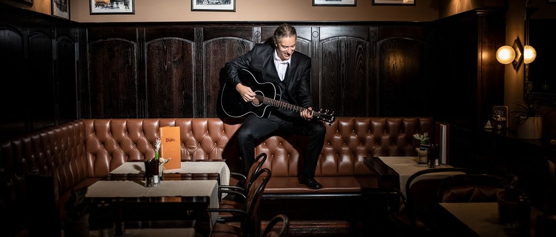 Ein Mann in einem Anzug spielt Gitarre auf einer Bank in einem Restaurant. Der Raum hat Holzwände, Holzkabinen und gerahmte Fotos. Es gibt Tische und Stühle aufgestellt.