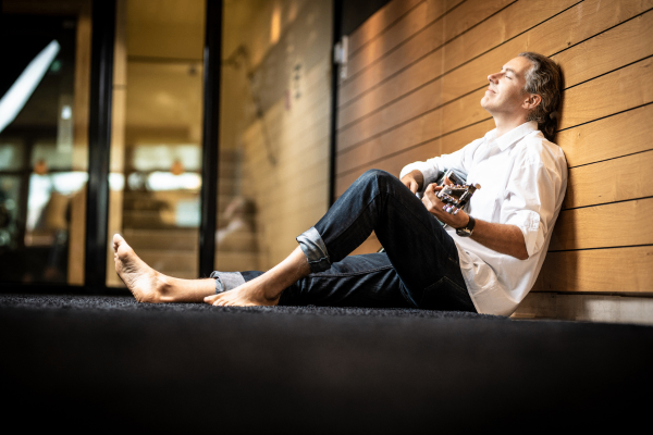 Ein Mann sitzt barfuß auf einer Bühne, mit geschlossenen Augen, und spielt eine Gitarre. Er lehnt sich gegen eine Holzwand.