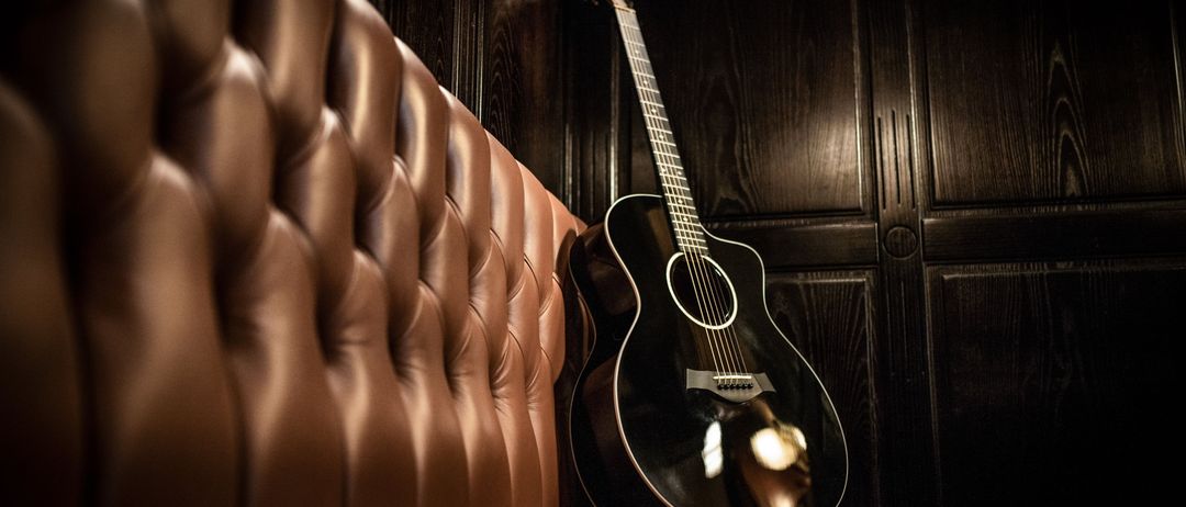 Eine schwarze Akustikgitarre liegt auf einem braunen Ledersofa vor einer dunklen Holzwand. Der glänzende Lack der Gitarre reflektiert das Licht und betont das komplizierte Design.